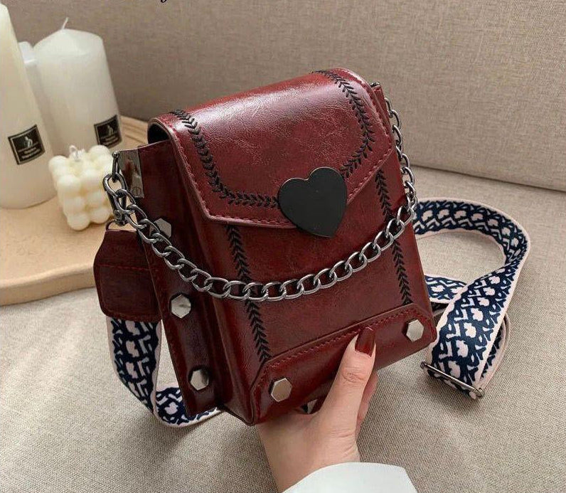 Brown leather bag with heart detail and floral strap held by a hand on a neutral background
