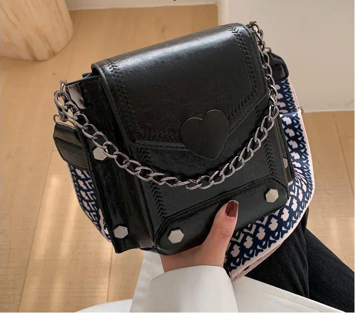 Black belt bag with heart detail and chain strap held by a person on a wooden floor.