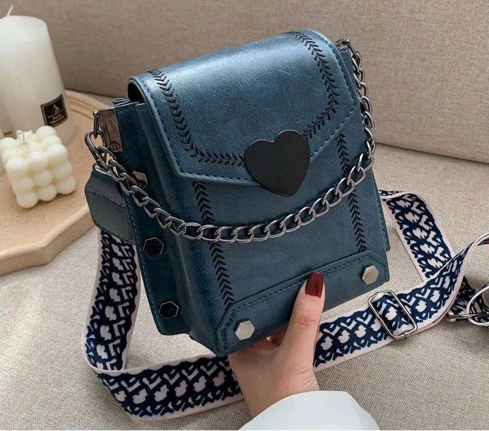 Blue handbag with heart detail and chain strap held by a hand on a neutral background