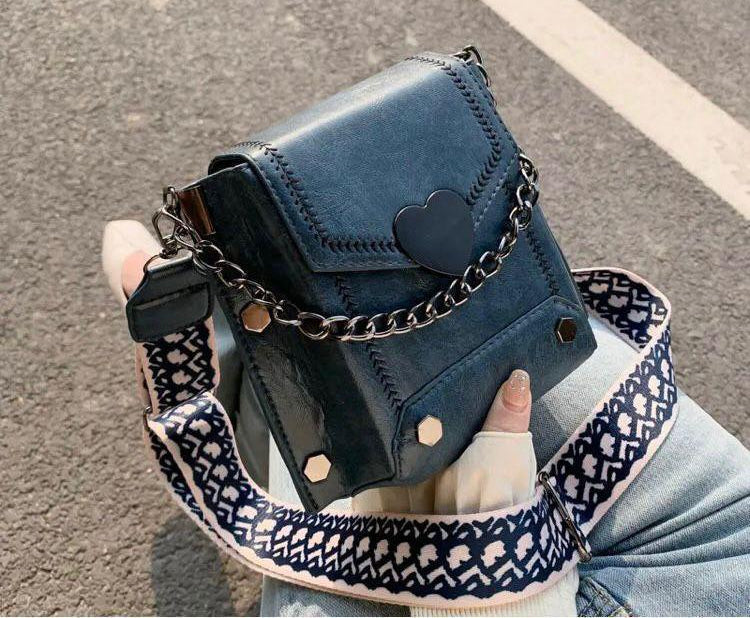 Blue crossbody bag with chain detail held by a person on a road background