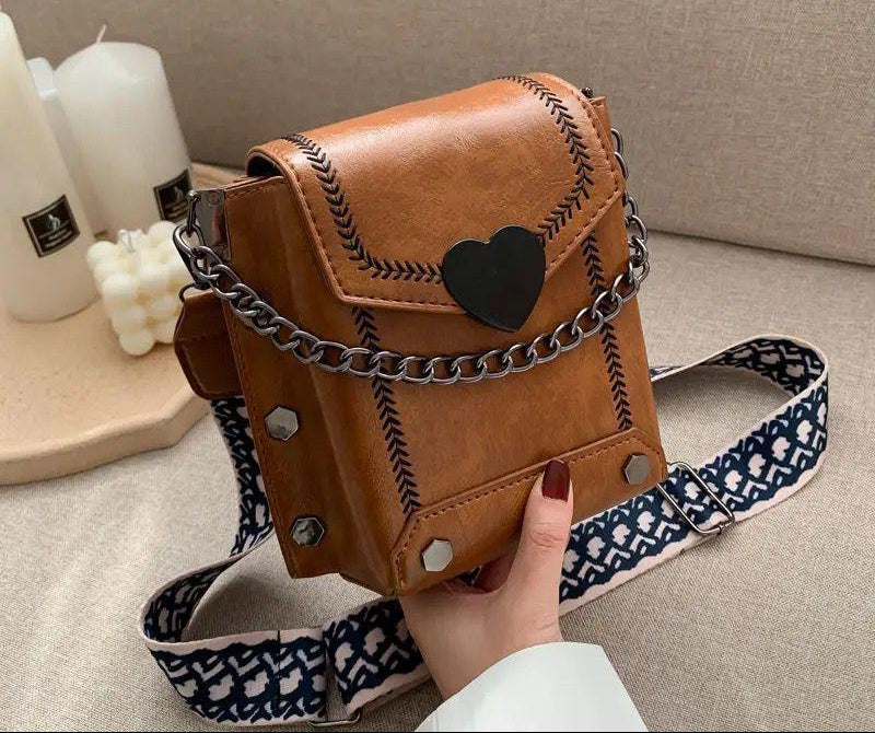 Brown leather bag with chain detail and patterned strap held by a hand on a neutral background.
