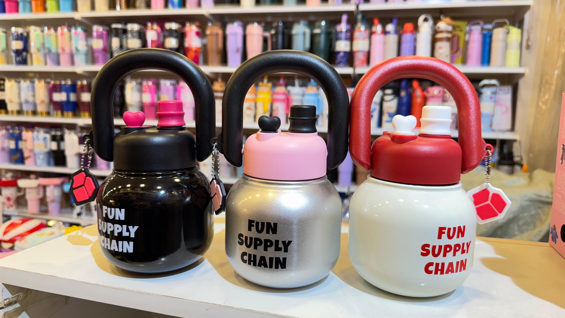 Three colorful water bottles shaped like kettlebells on a shelf with stationery items in the background.