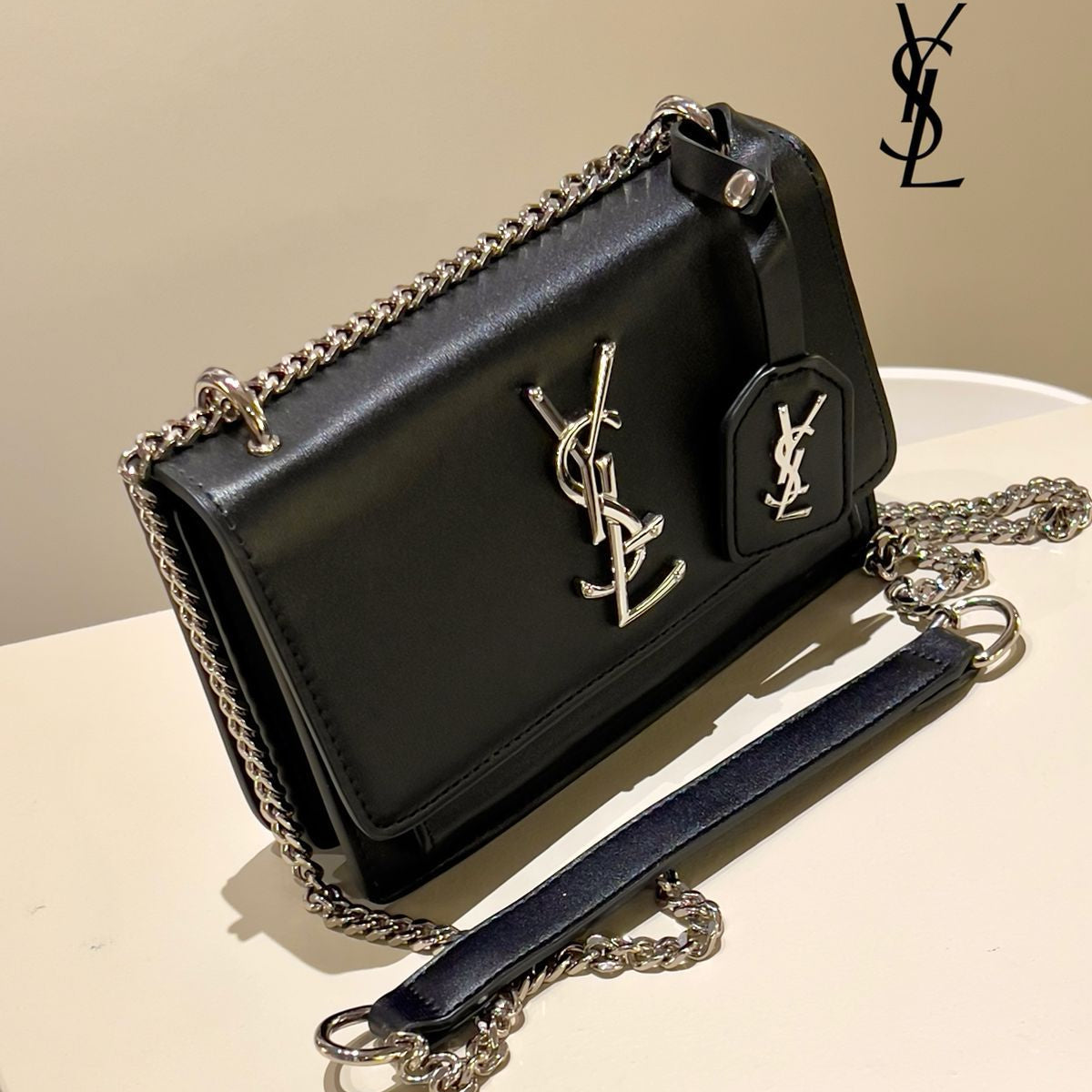 Black handbag with chain detail on a beige surface, featuring Yves Saint Laurent logo.