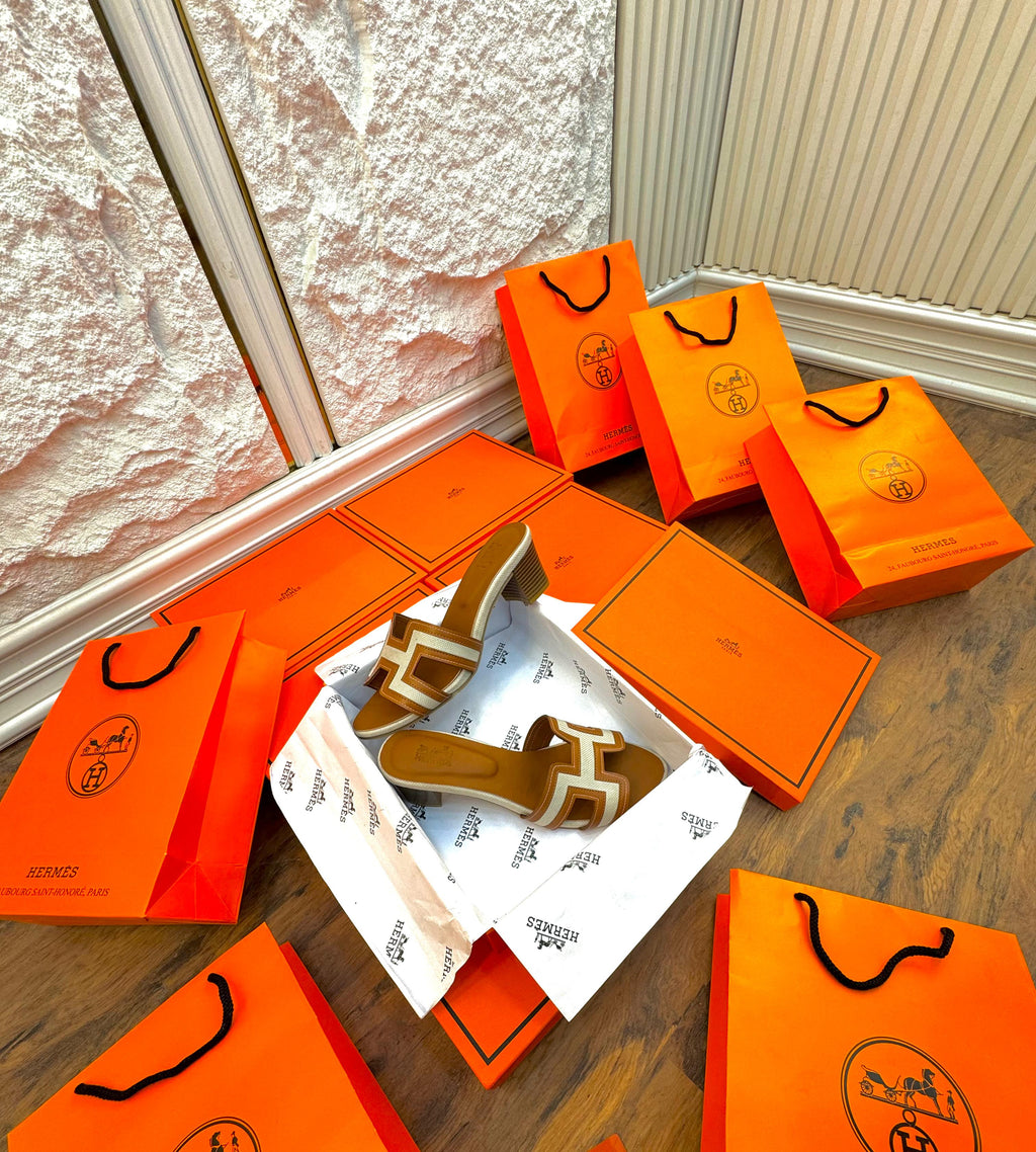 Brown HERMES Heel sandals with white soles in an open orange shoe box, surrounded by multiple closed orange shoe boxes on a wooden floor.
