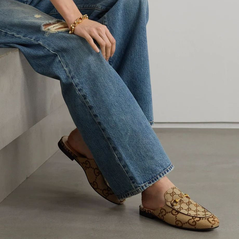 Brown Gucci mules  wearing blue jeans and patterned loafers sitting on a concrete surface.