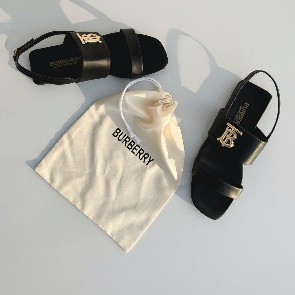 Black sandals with gold logo on a marble surface, accompanied by a Burberry dust bag.