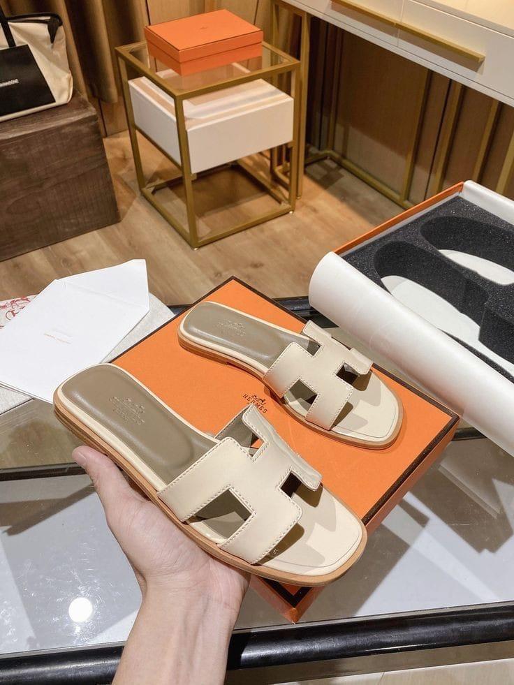 Beige HERMES Oran Velvet women  sandal held over an orange Hermes box with a blurred indoor setting.