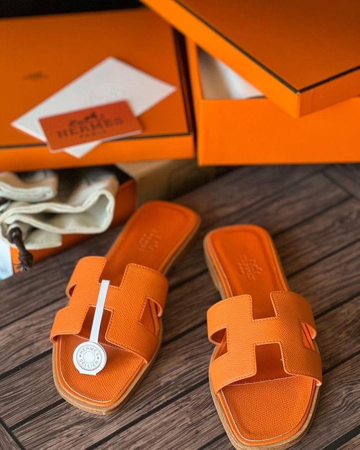 Orange HERMES Oran Velvet women  sandals with a brand logo on a wooden surface, with an orange box in the background.