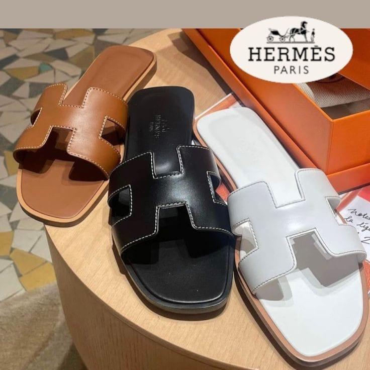 Three Hermès sandals in brown, black, and white on a wooden surface with an orange box in the background.