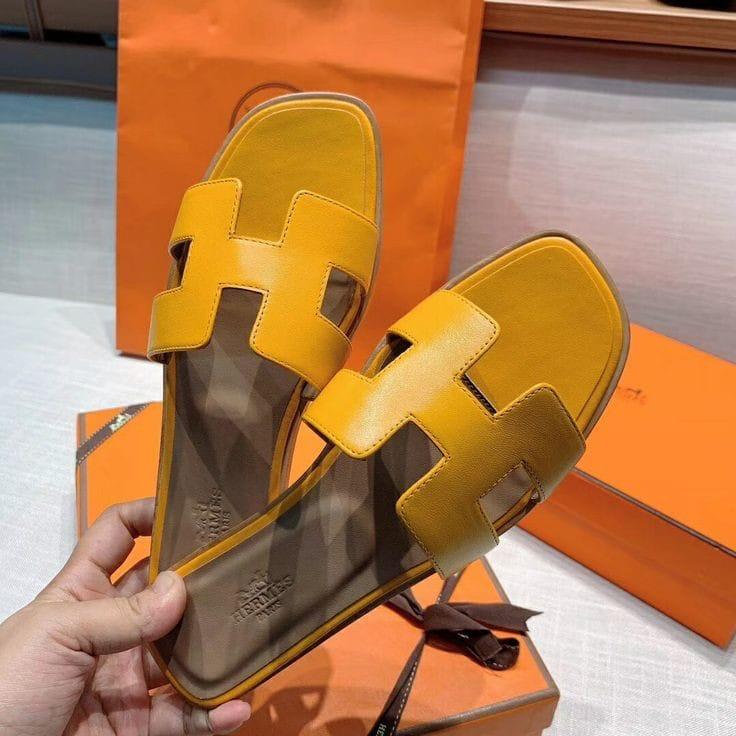 Yellow White HERMES Oran Velvet women heels sandals with a brand logo held by a hand, placed on orange boxes.