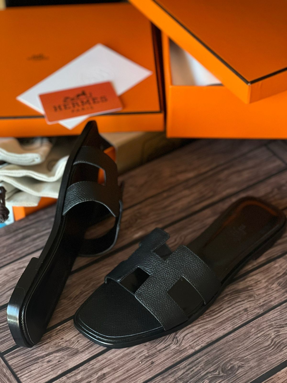 Black HERMES Oran Velvet women  sandals on a wooden surface with an orange Hermes box in the background.