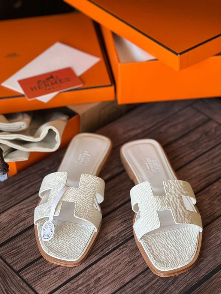 White HERMES Oran Velvet women   sandals with a brand logo on a wooden surface, with an orange shoe box in the background.