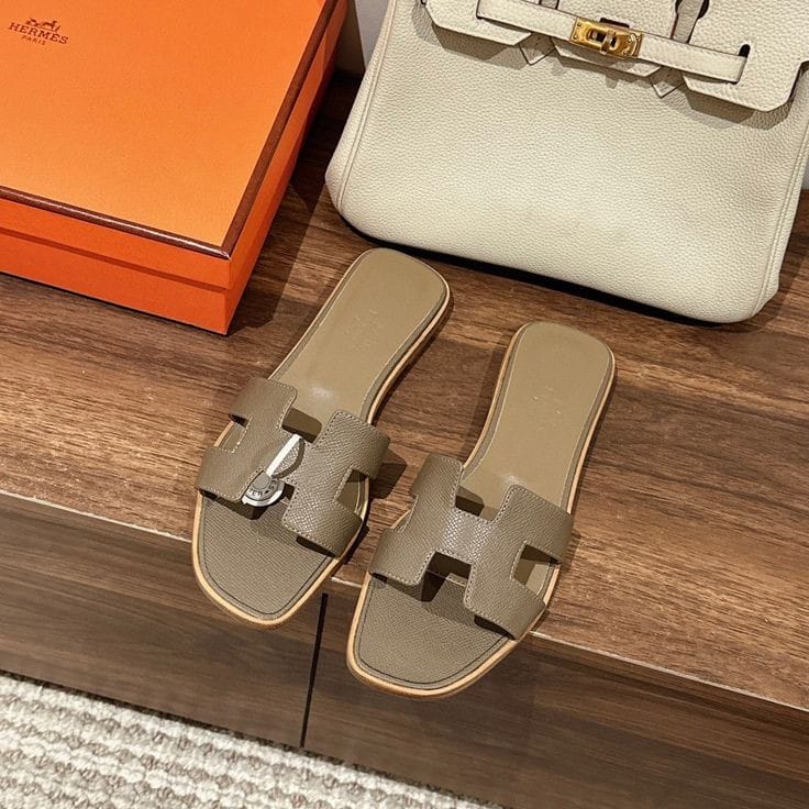 Gold HERMES Oran Velvet women  sandals with a visible brand logo on a wooden surface, next to an orange box and a white handbag.
