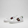 White Gucci Ace Embroidered ‘Bee’   sneakers with colorful stripes worn by a person on a wooden deck.