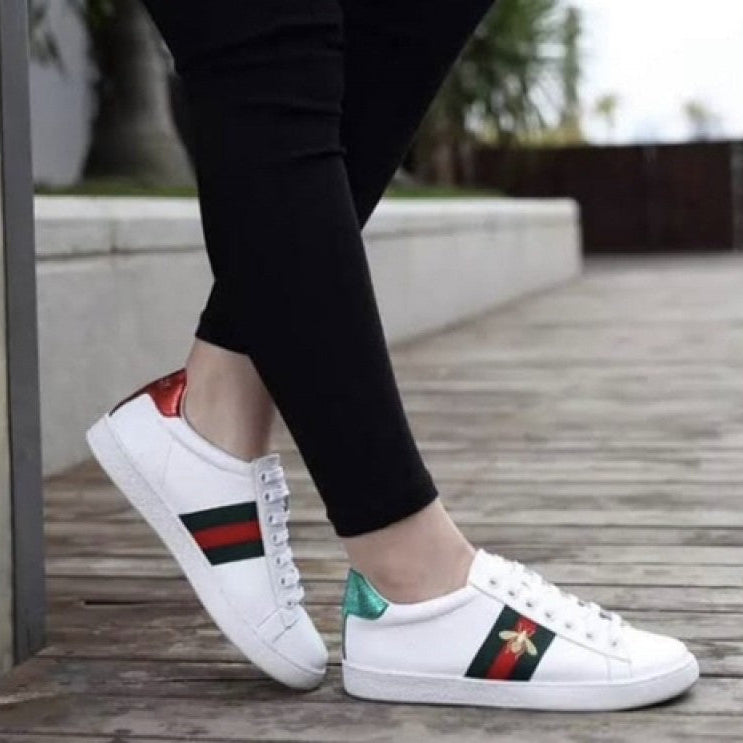White Gucci Ace Embroidered ‘Bee’   sneakers with colorful stripes worn by a person on a wooden floor.