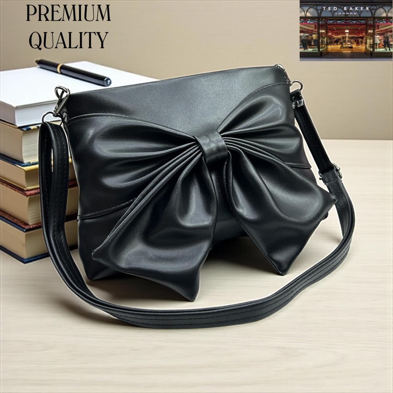 Black TED BAKER London Premium Sling Bag handbag with a large bow on a beige surface, with books and a pen in the background.