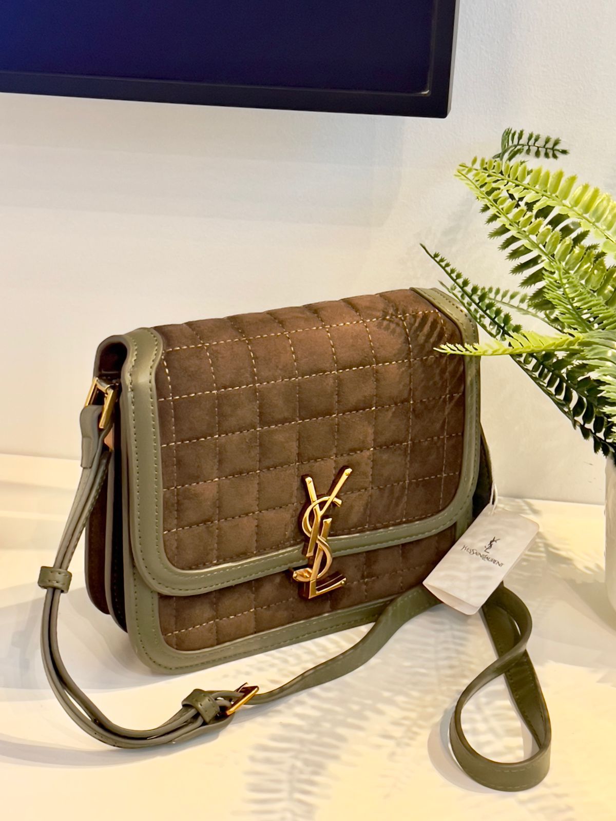 Tan Yves Saint Laurent YSL Premium Shoulder Bag quilted handbag with green strap on a light surface with a plant in the background