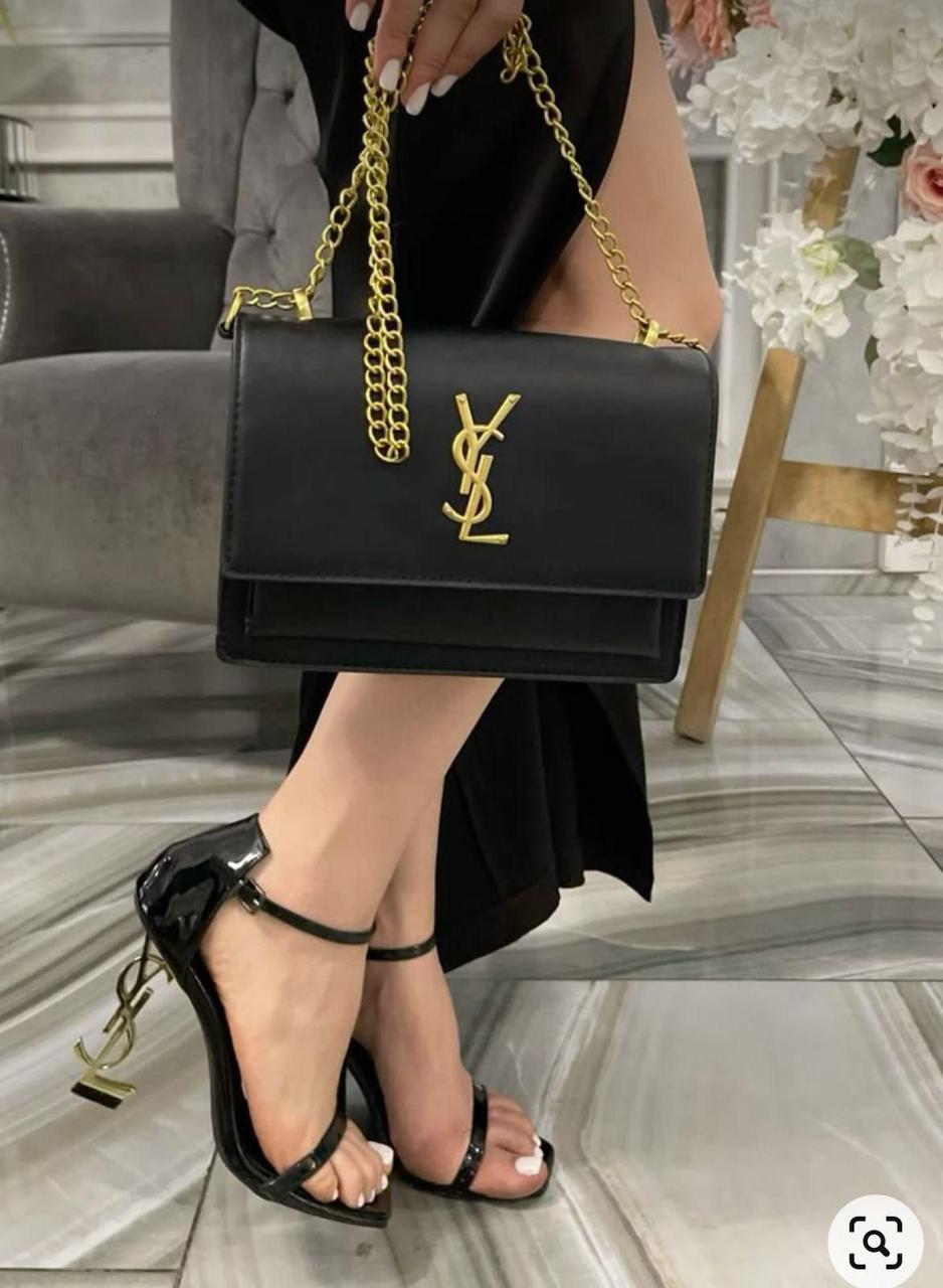 YSL High Heel Sandals for Women Person holding a black handbag with a gold chain strap, wearing black high-heeled sandals.
