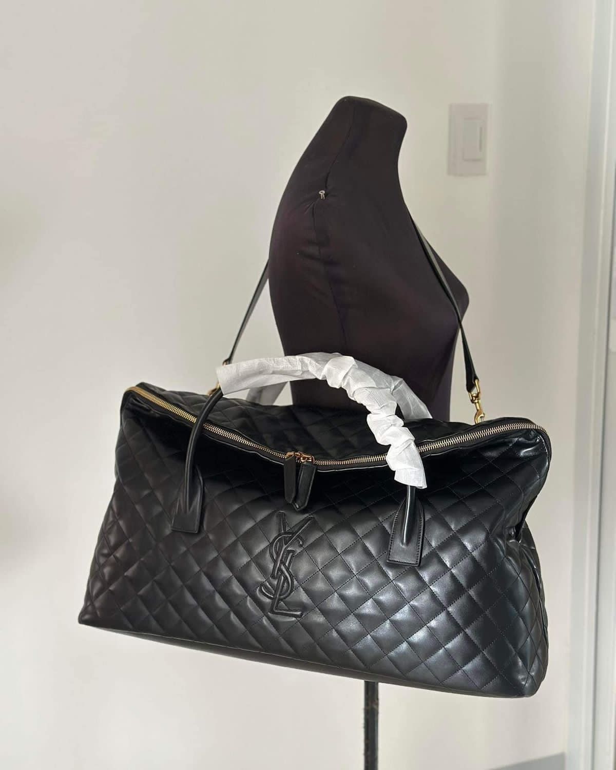 Black YSL Giant Quilted Leather Weekend Duffle Bag Black quilted handbag with brand logo on a mannequin against a white wall.