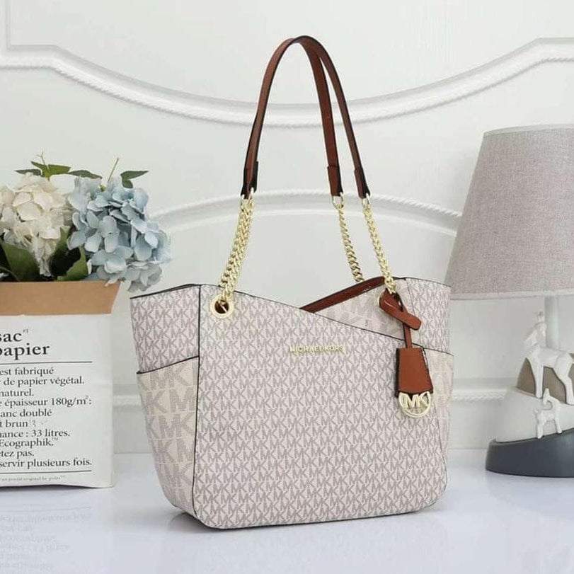 White Michael Kors Signature Tote Bag handbag with brown handles and strap on a white surface with a lamp and flowers in the background.