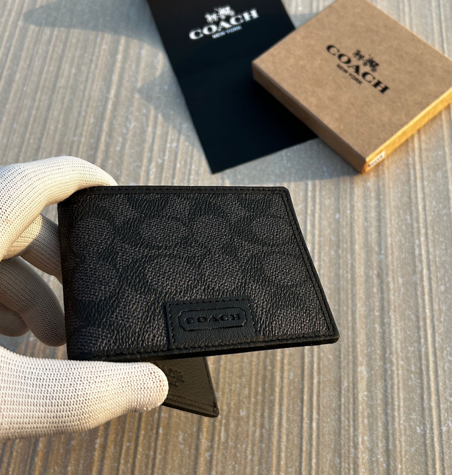 Black leather wallet with Coach logo held by a gloved hand, with Coach packaging in the background.