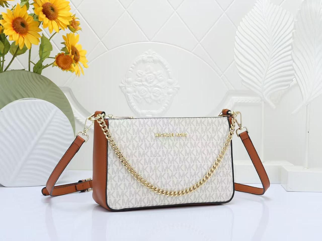 Brown Michael Kors Chain Sling Bag handbag with brown accents and gold chain strap on a white surface with sunflowers in the background.
