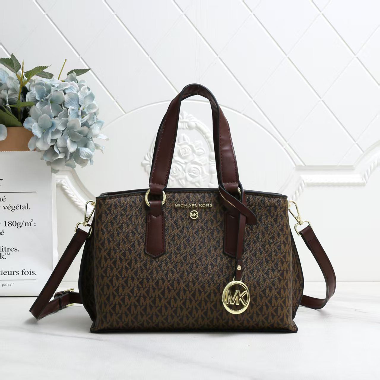 Tan Michael Kors Marilyn Medium Satchel Brown handbag with gold accents on a white surface with a light gray textured wall background