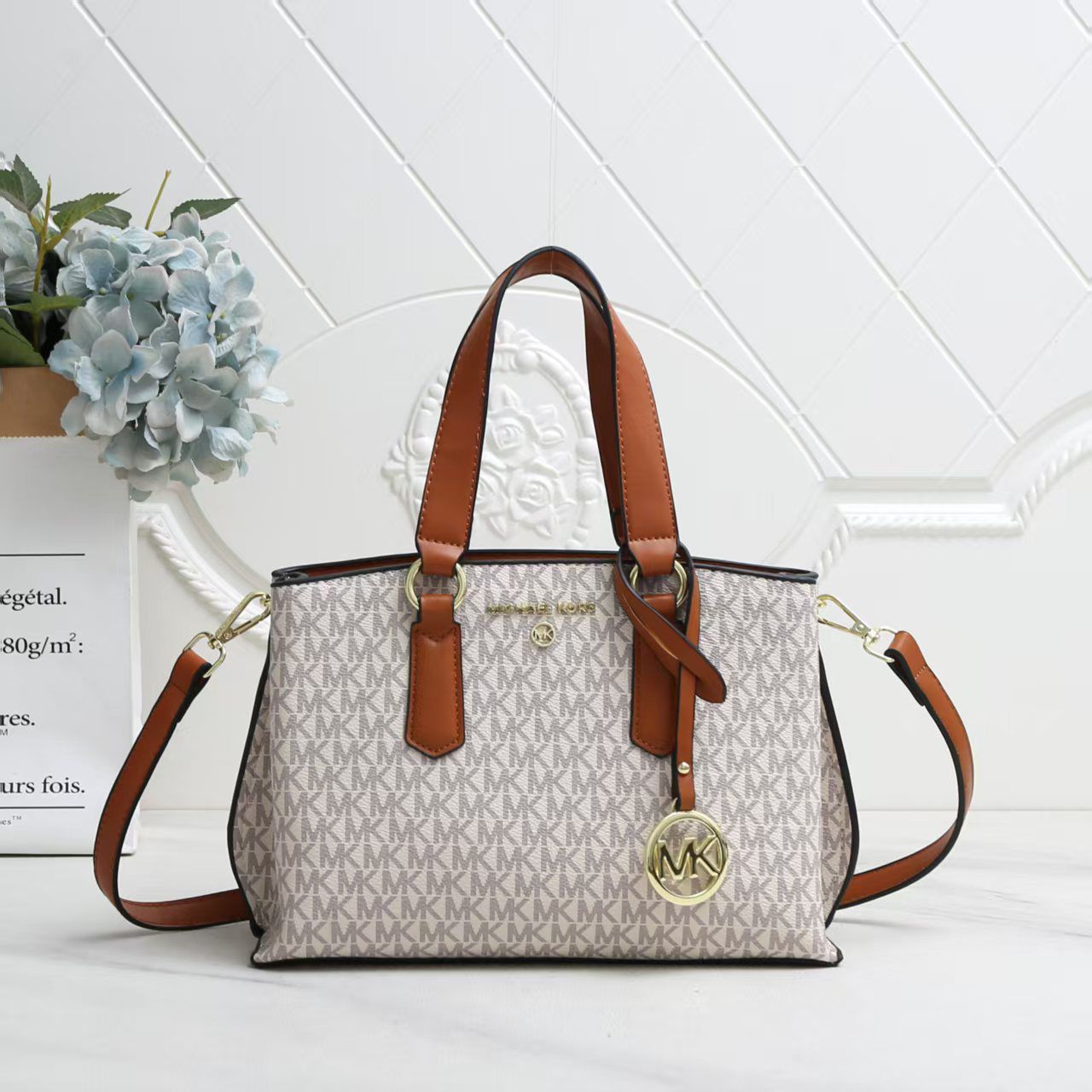 White Michael Kors Marilyn Medium Satchel Designer handbag with brown straps on a white surface with a light gray geometric patterned wall in the background.