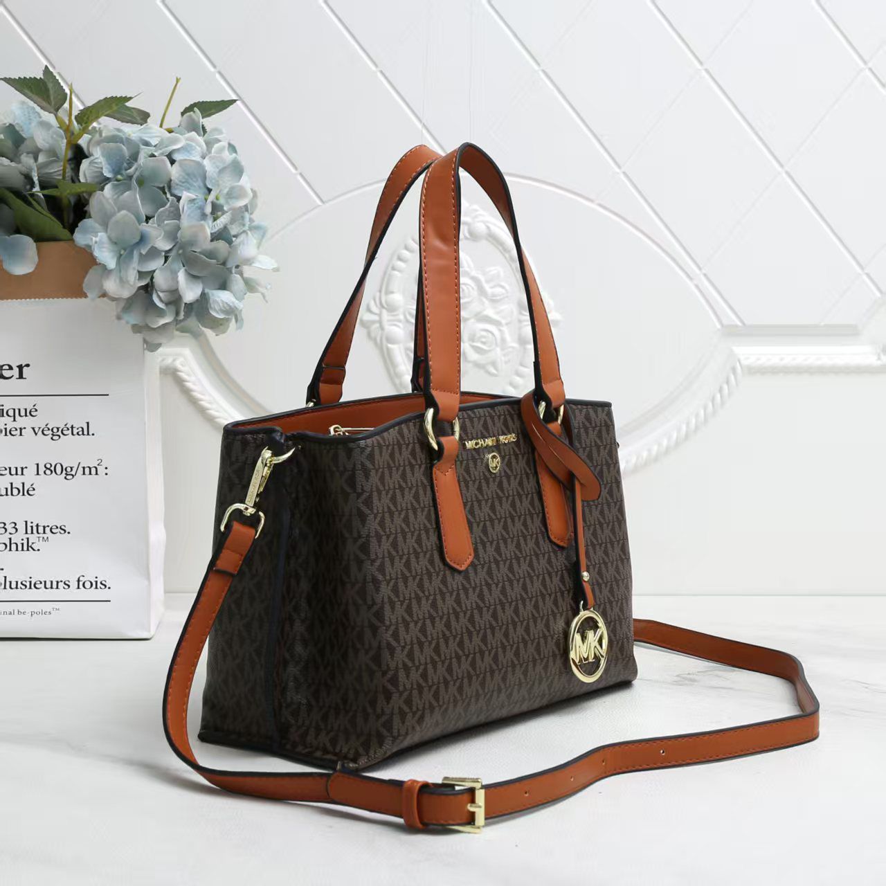 Tan Michael Kors Marilyn Medium Satchel Brown handbag with gold accents on a white surface with a light blue floral arrangement in the background.