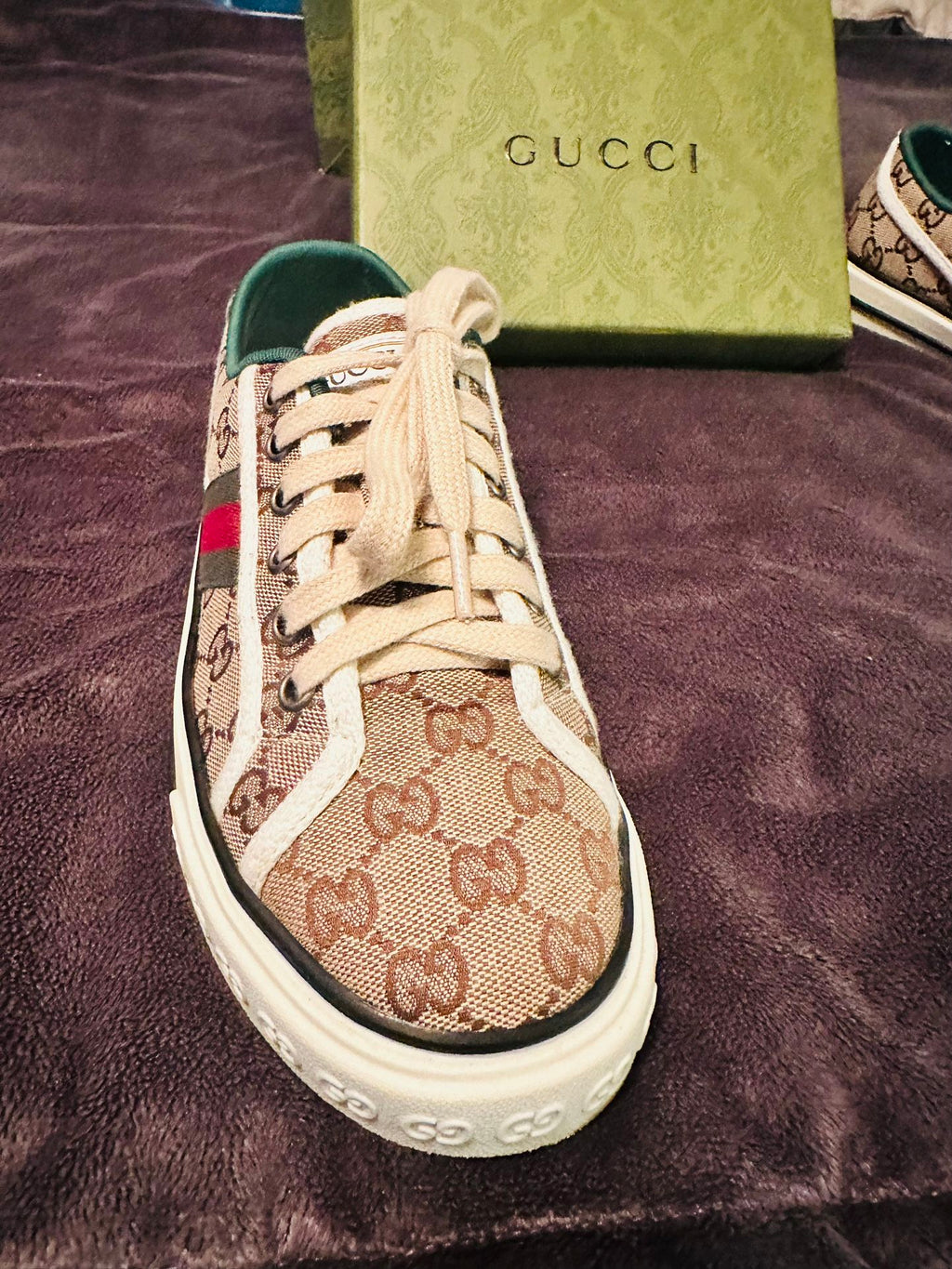 Gucci sneaker with patterned design on a purple surface, Gucci box visible.