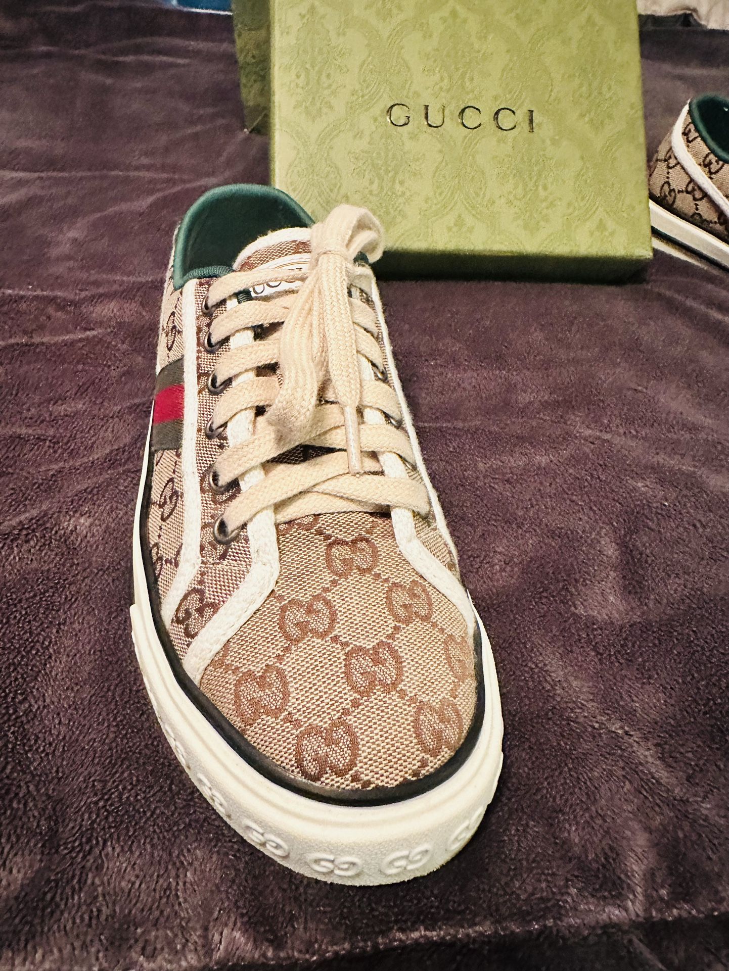 Gucci sneaker with patterned design on a purple surface, Gucci box visible.