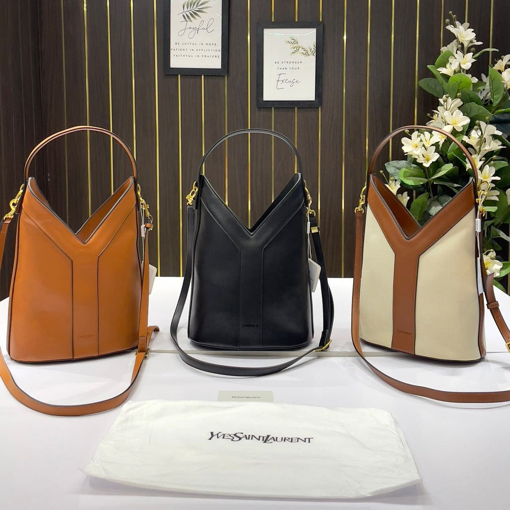 YSL Y Top Handle Bucket Bag Three leather handbags in brown, black, and beige with a YSL pouch on a striped wall background.